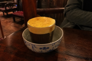 Glass of Hanoi egg coffee with thick yellow foam sitting in a bowl of hot water