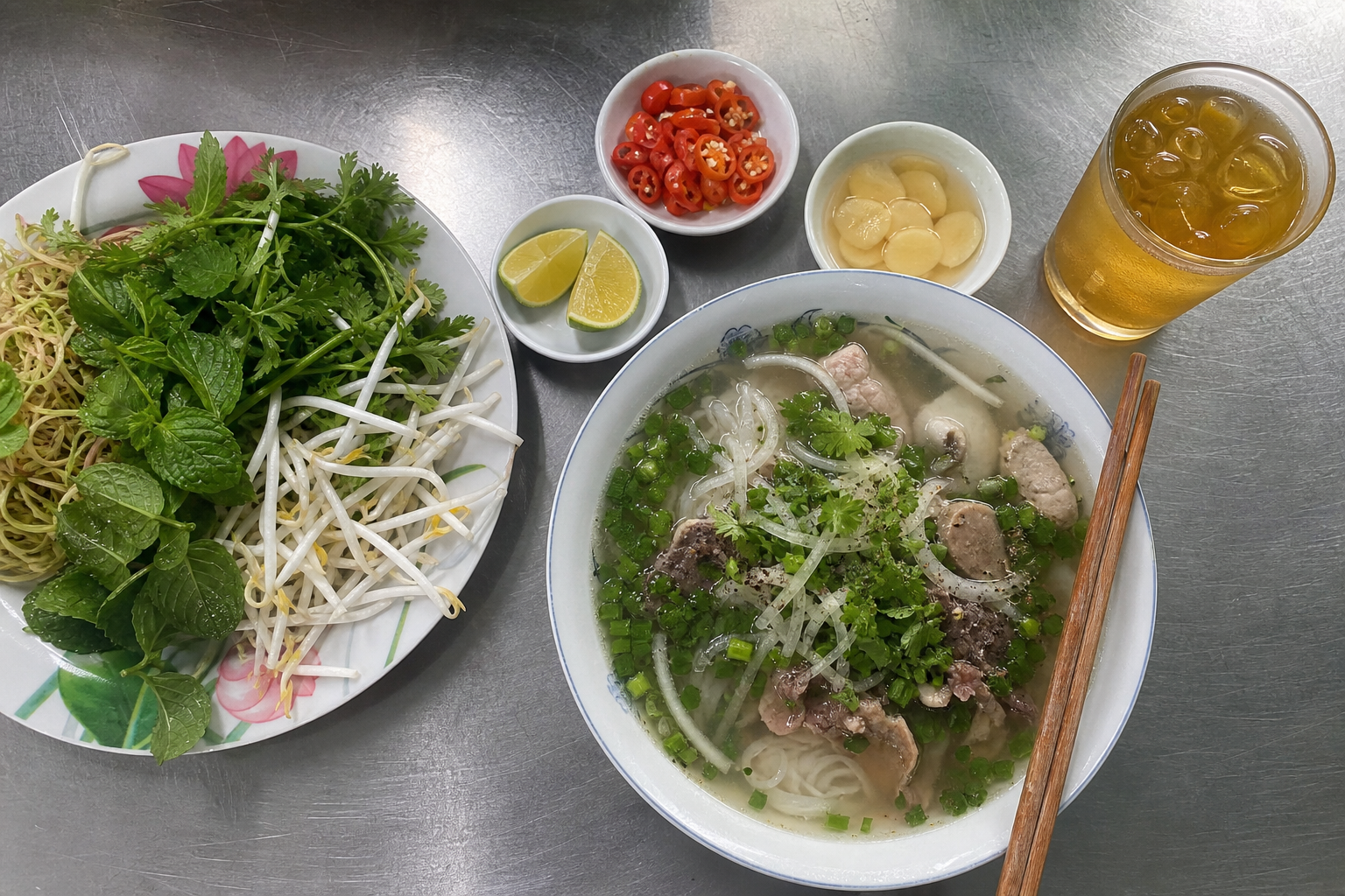 Bowl of Vietnamese noodle soup with herb plate, chili, lime and garlic vinegar on a steel table
