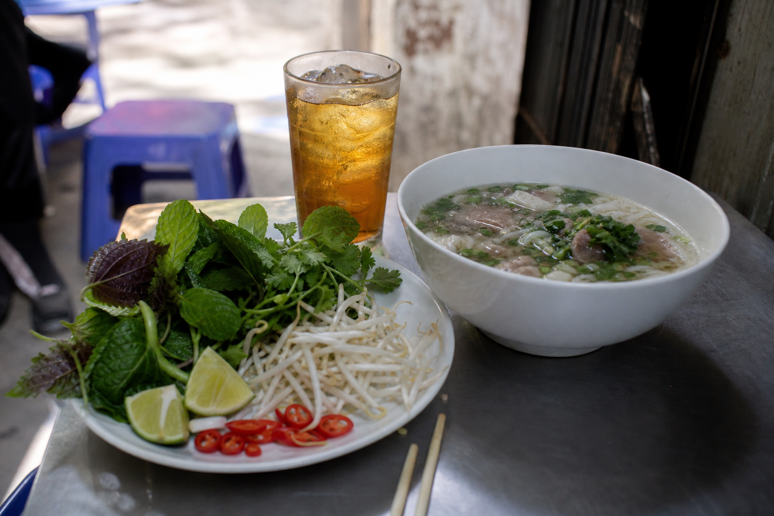 Plate of fresh Vietnamese herbs with lime and chilli beside a bowl of noodle soup and a glass of iced tea on a low street-side table