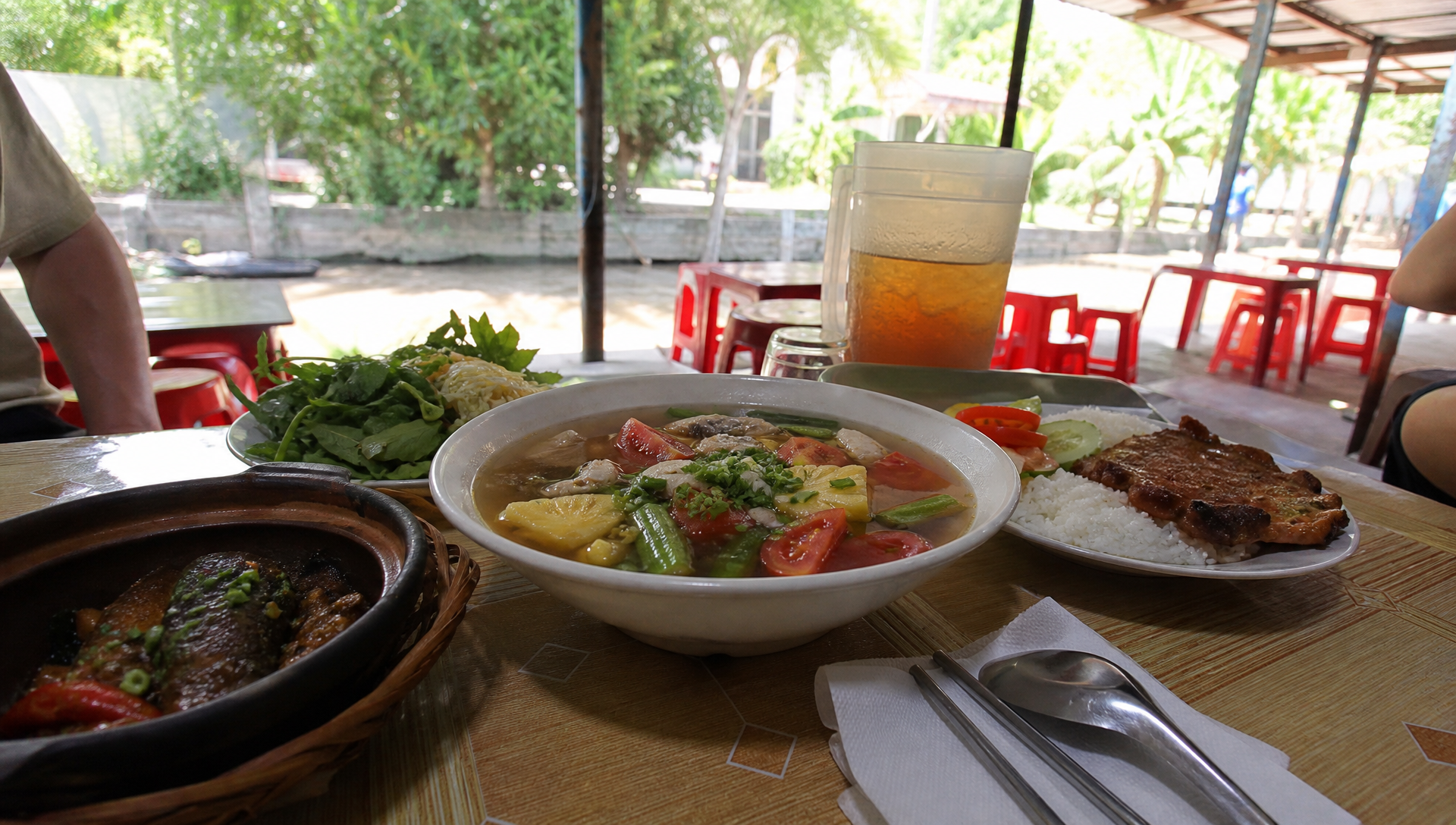 Roadside Mekong Delta eatery table with a bowl of sour soup, rice, grilled pork and a clay pot of braised fish