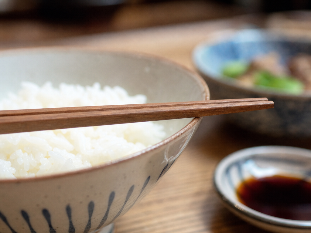 Chopsticks resting correctly across the rim of a ceramic rice bowl rather than standing upright in the rice