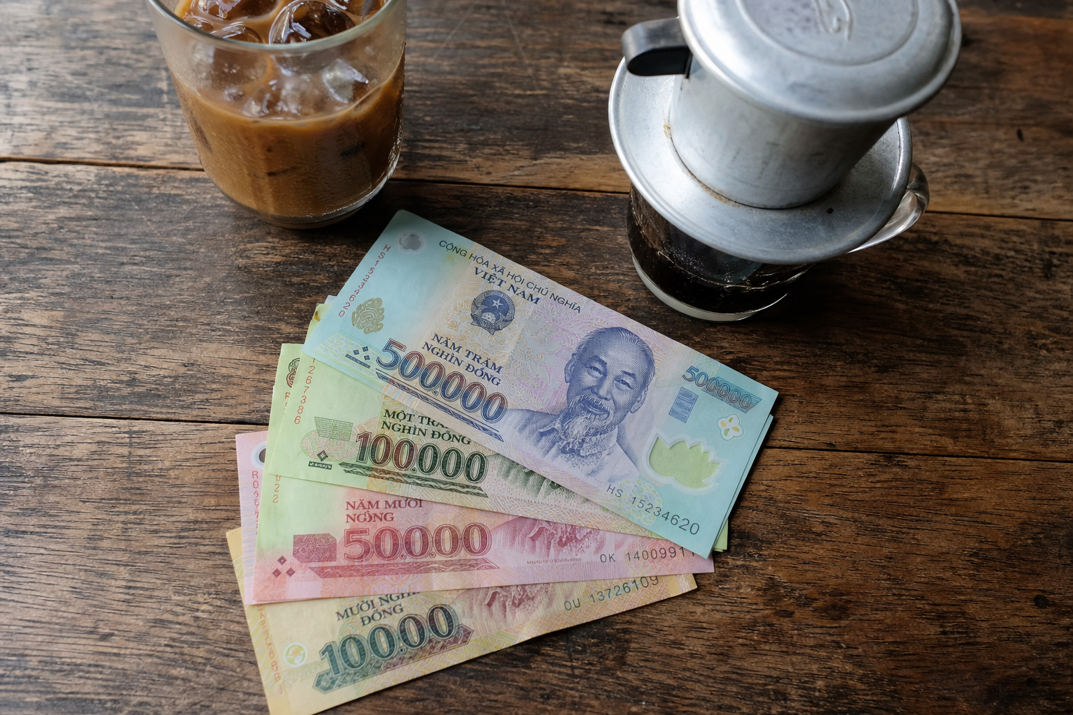 Vietnamese dong polymer banknotes fanned out on a wooden cafe table beside an iced coffee and phin filter