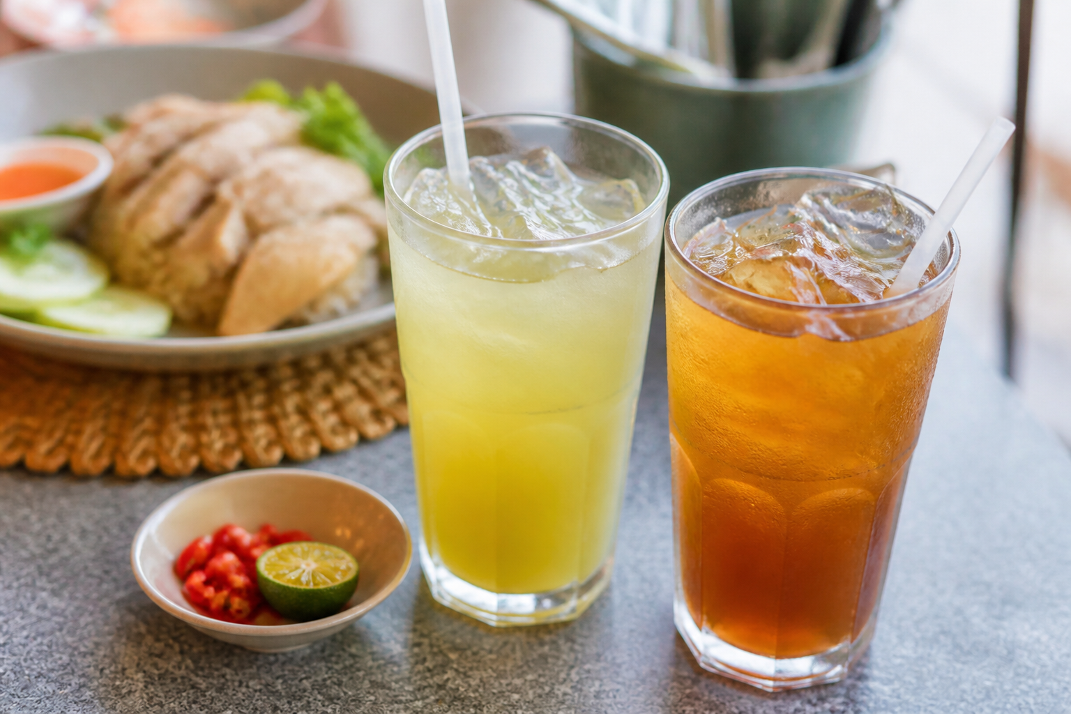 Glass of sugarcane juice and iced tea with sliced chilli and lime wedge beside a bowl of chicken rice